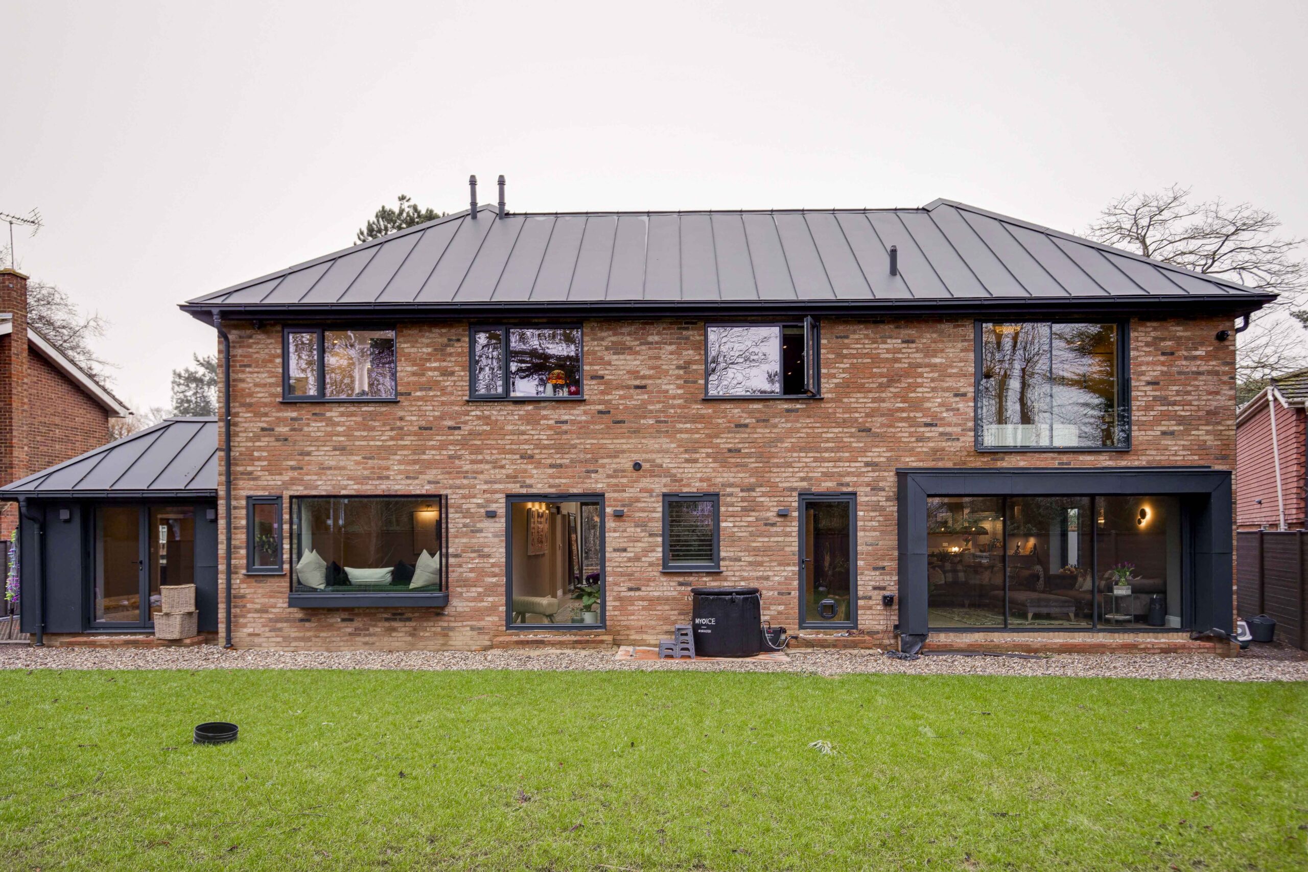 The garden elevation at Woodfield Road showing brick, standing seam metal roof and large format aluminium glazing across the extension and the rebuilt house